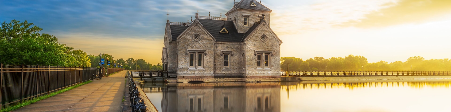 A historic stone building with a reflection in calm water, surrounded by a fence and trees.