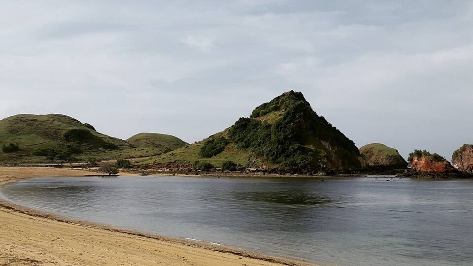 A beach with a calm sea, green hills, and a prominent hill in the background.