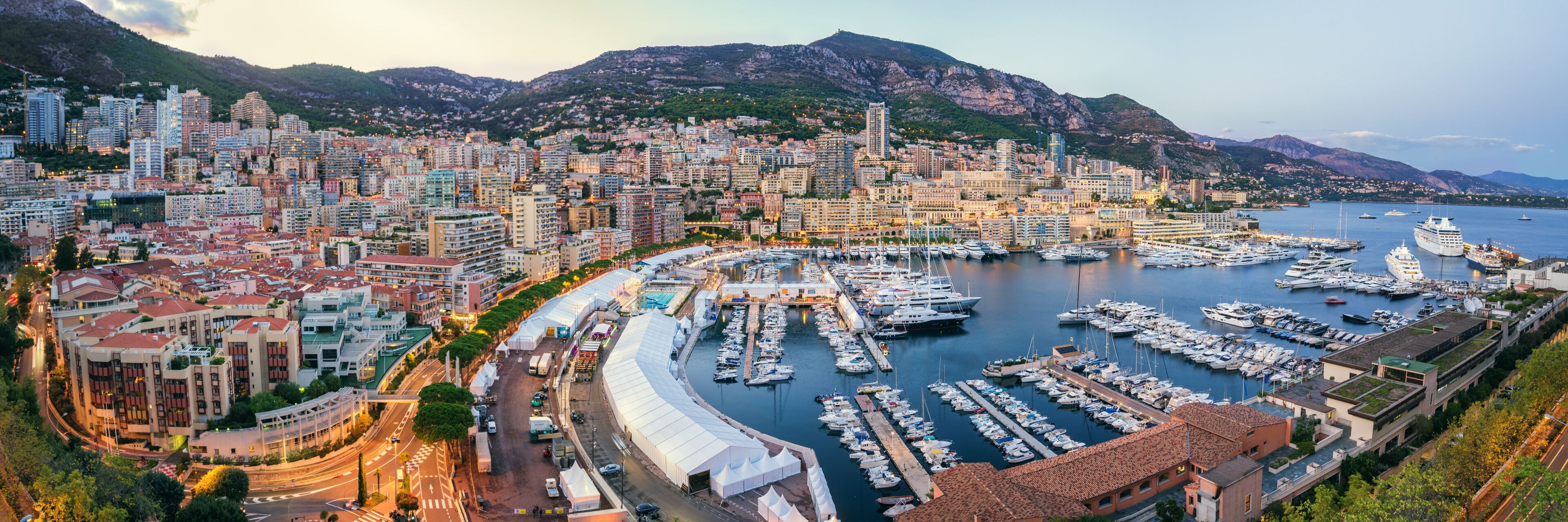 Paesaggio costiero con una marina piena di barche, circondato da montagne e edifici urbani.