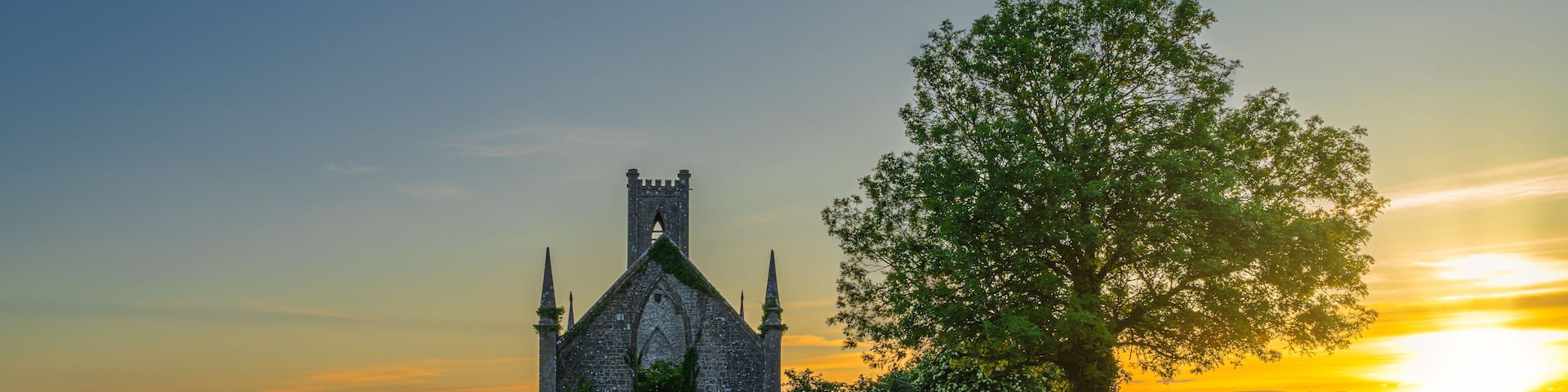Cereal field illuminated by sunlight and old stone ruins of Ballinafagh Church with dramatic sky at sunset in background, County Kildare, Ireland