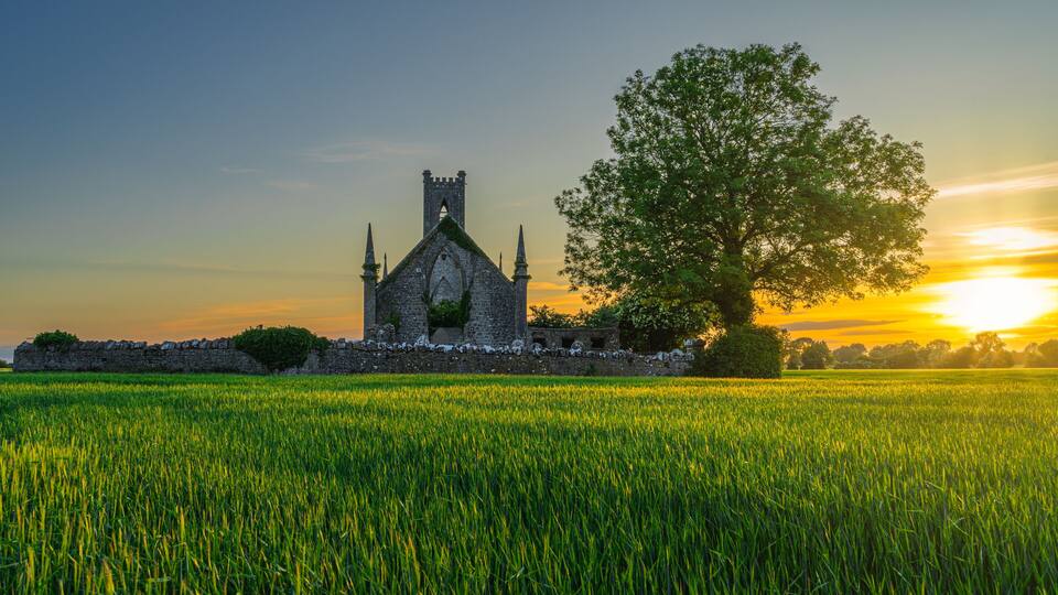 A medieval stone church with a large tree in front, set against a sunset sky.