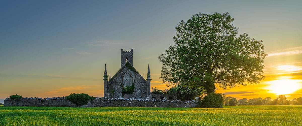 A medieval stone church with a large tree in front, set against a sunset sky.