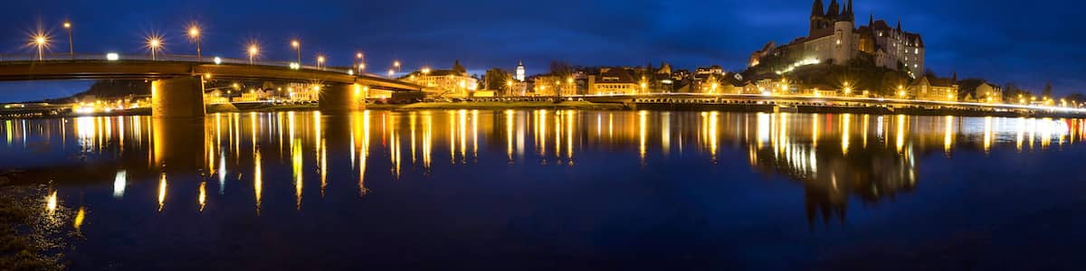 A nighttime view of a city with a prominent castle on a hill, a bridge spanning a river, and city lights reflecting on the water.