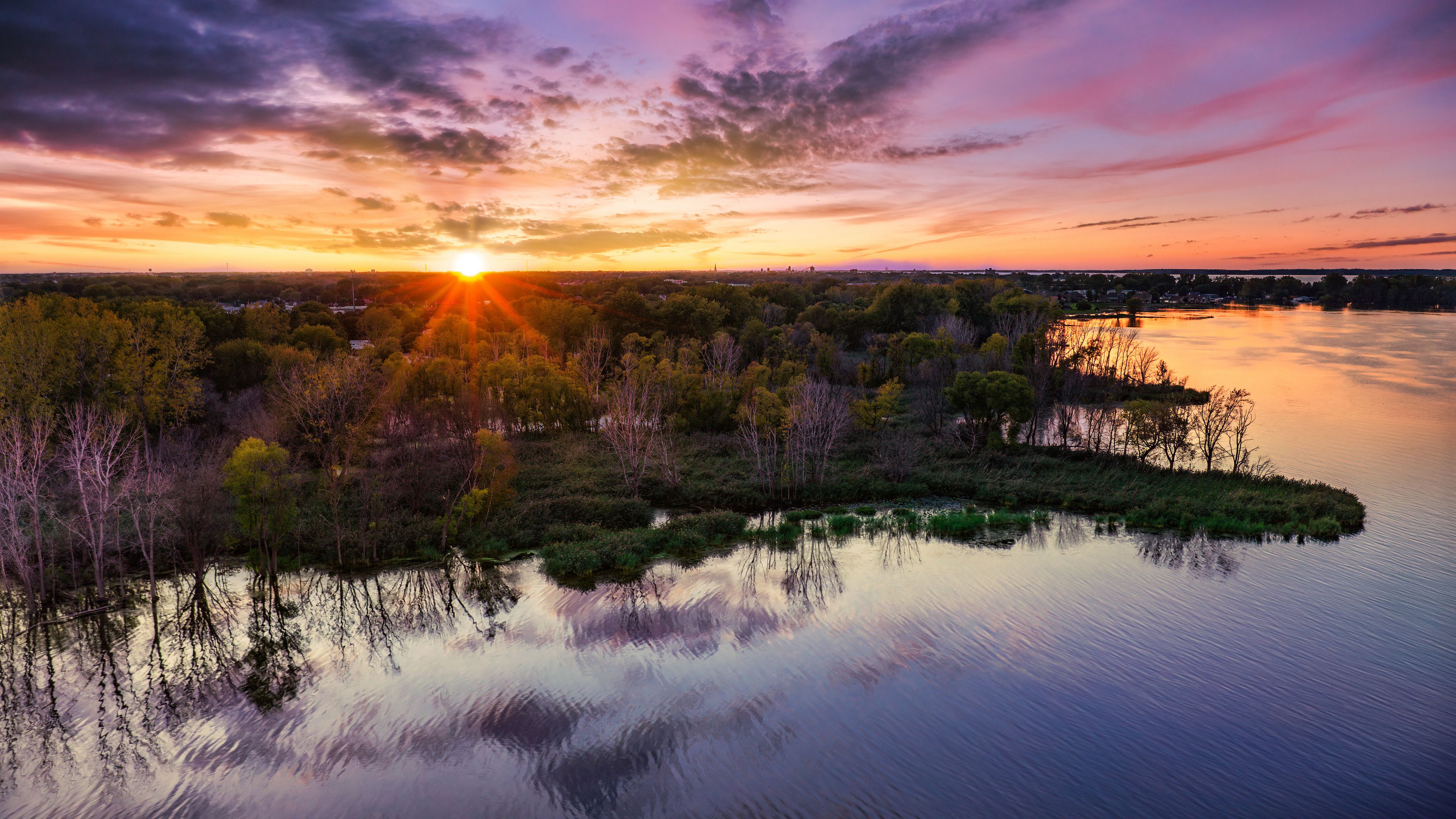 A sunset over a calm lake with trees reflecting the sky, surrounded by a forested area.
