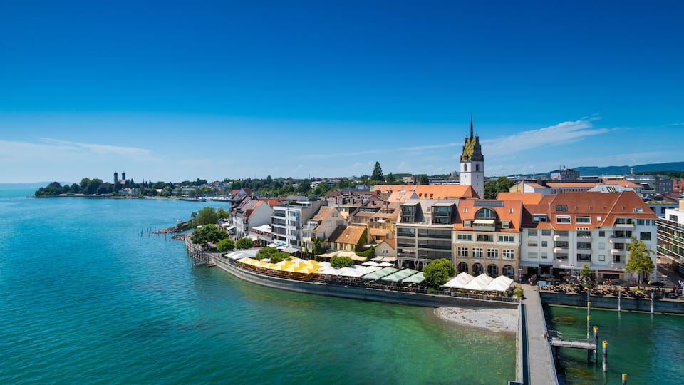 A coastal town with a prominent church tower, waterfront cafes, and a clear blue sky.