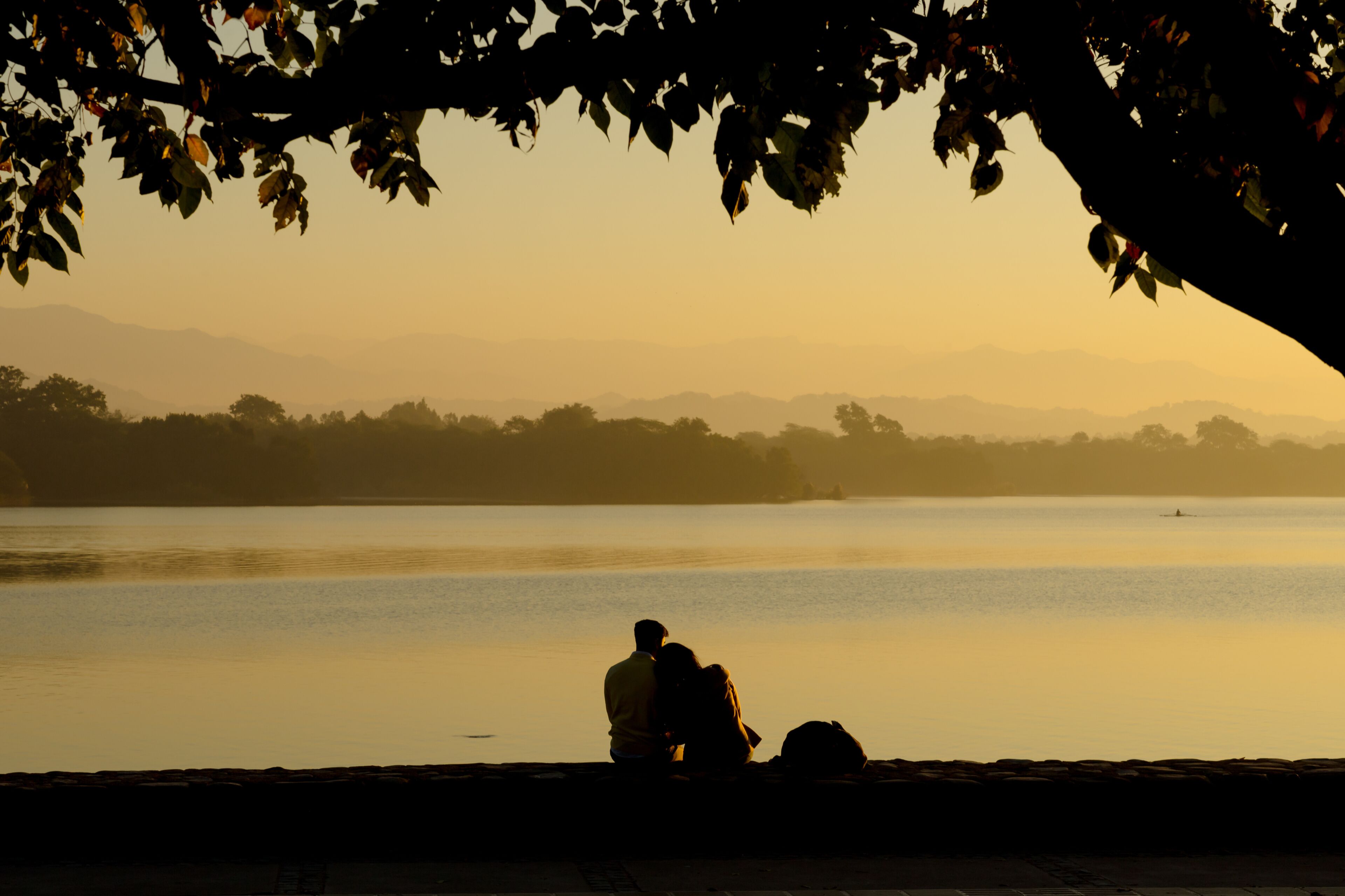 Um casal sentado à beira de um lago calmo durante o pôr do sol, com montanhas ao fundo.