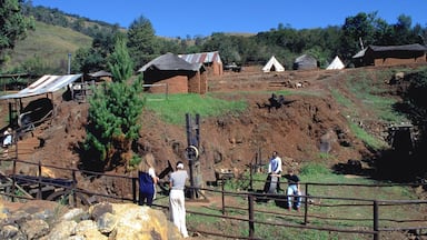 Paisagem rural com uma colina de terra, alguns edifícios pequenos e uma cerca.