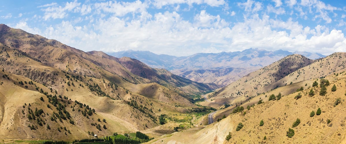 A mountainous landscape with a valley, scattered trees, and distant mountains under a partly cloudy sky.