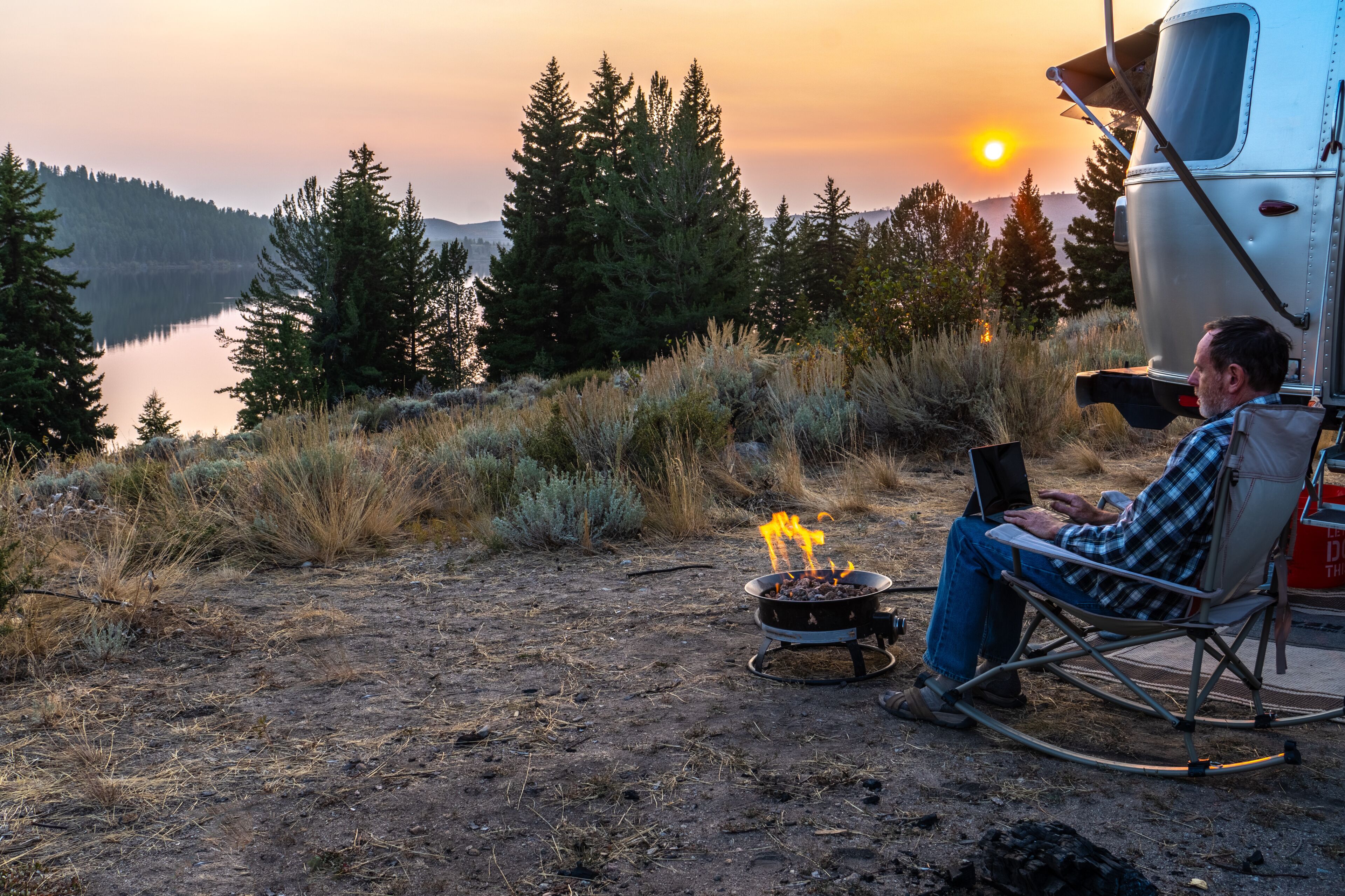 Person sitting on a rocking chair by the side of a travel trailer, RV, campfire working on a laptop, PC as the sun setting over a distance hill, mountain, Half Moon Lake, Pinedale, Wyoming