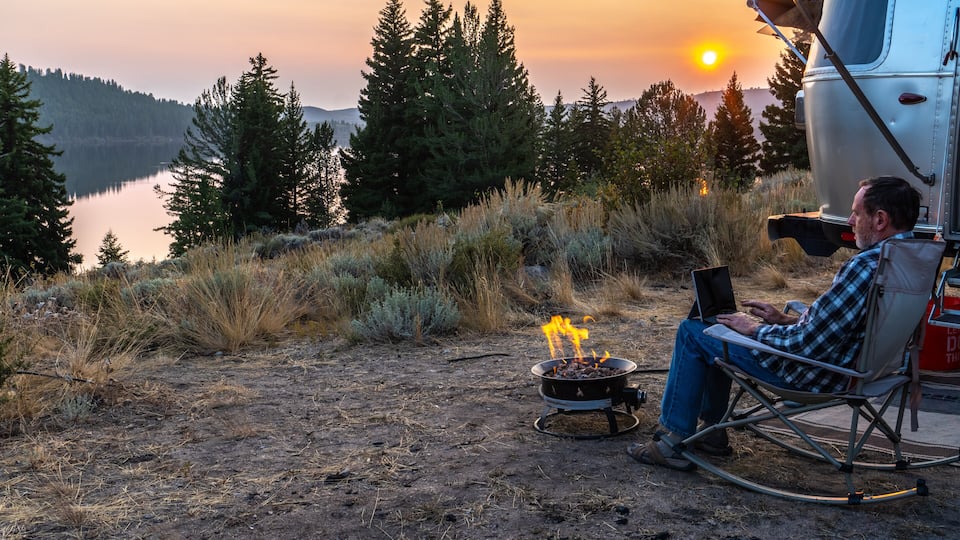 A man in a rocking chair with a laptop, a campfire, and a camper by a lake at sunset.