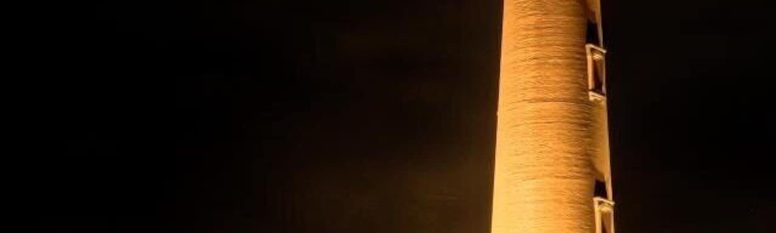 Um farol alto e iluminado à noite, com uma pequena casa nas proximidades.