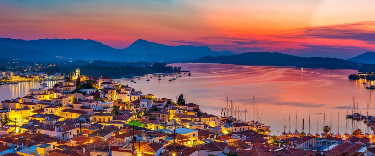 A coastal town at sunset with illuminated buildings, a calm sea, and mountains in the background.