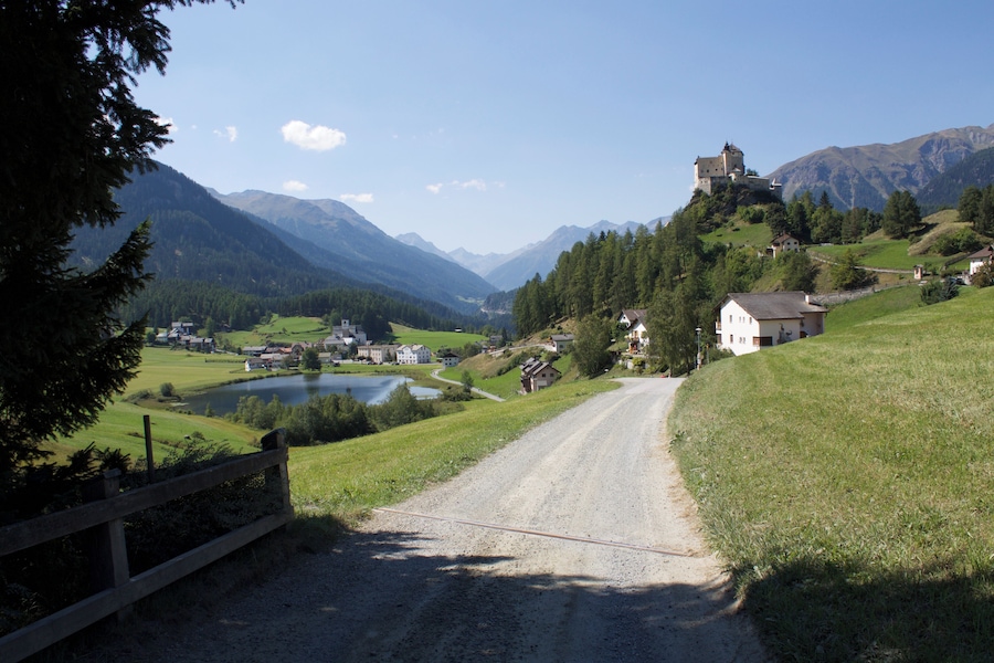 Ländliche Landschaft mit Feldweg, See, Häusern und Burg auf einem Hügel.