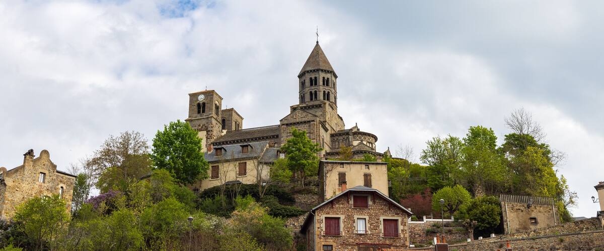 A historic stone building with a clock tower and a bell tower, surrounded by lush greenery and other stone structures.