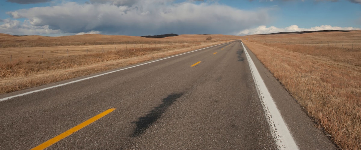 A straight road with yellow center lines, surrounded by dry grassland and a clear blue sky with large clouds.