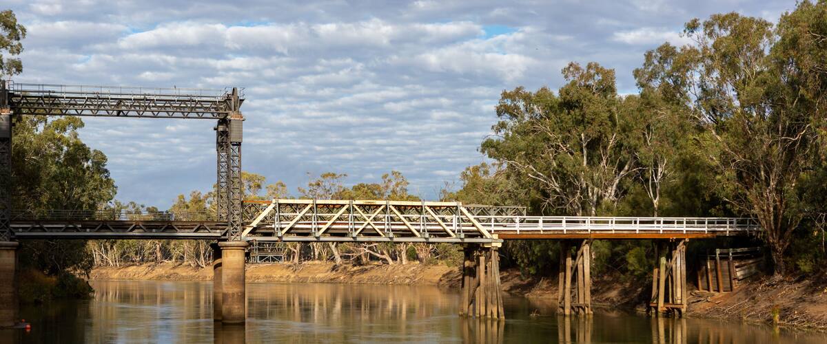 A steel bridge over a calm river with trees on either side.
