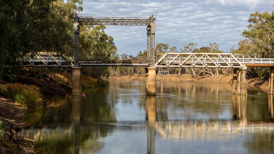 A metal bridge over a calm river, surrounded by trees and a cloudy sky.