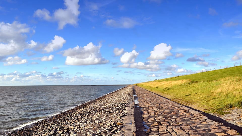 A coastal path with a rocky shoreline, a grassy hill, and a clear blue sky with clouds.