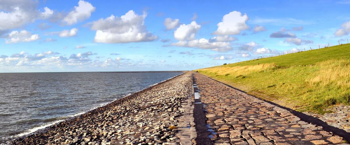 A coastal path with a rocky shoreline, a grassy hill, and a clear blue sky with clouds.