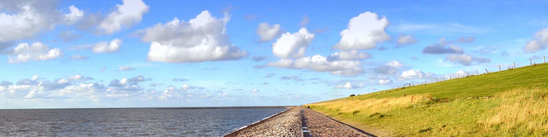A coastal path with a rocky shoreline, a grassy hill, and a clear blue sky with clouds.