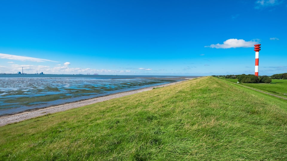 A coastal landscape with a lighthouse, grassy shore, and distant industrial structures.