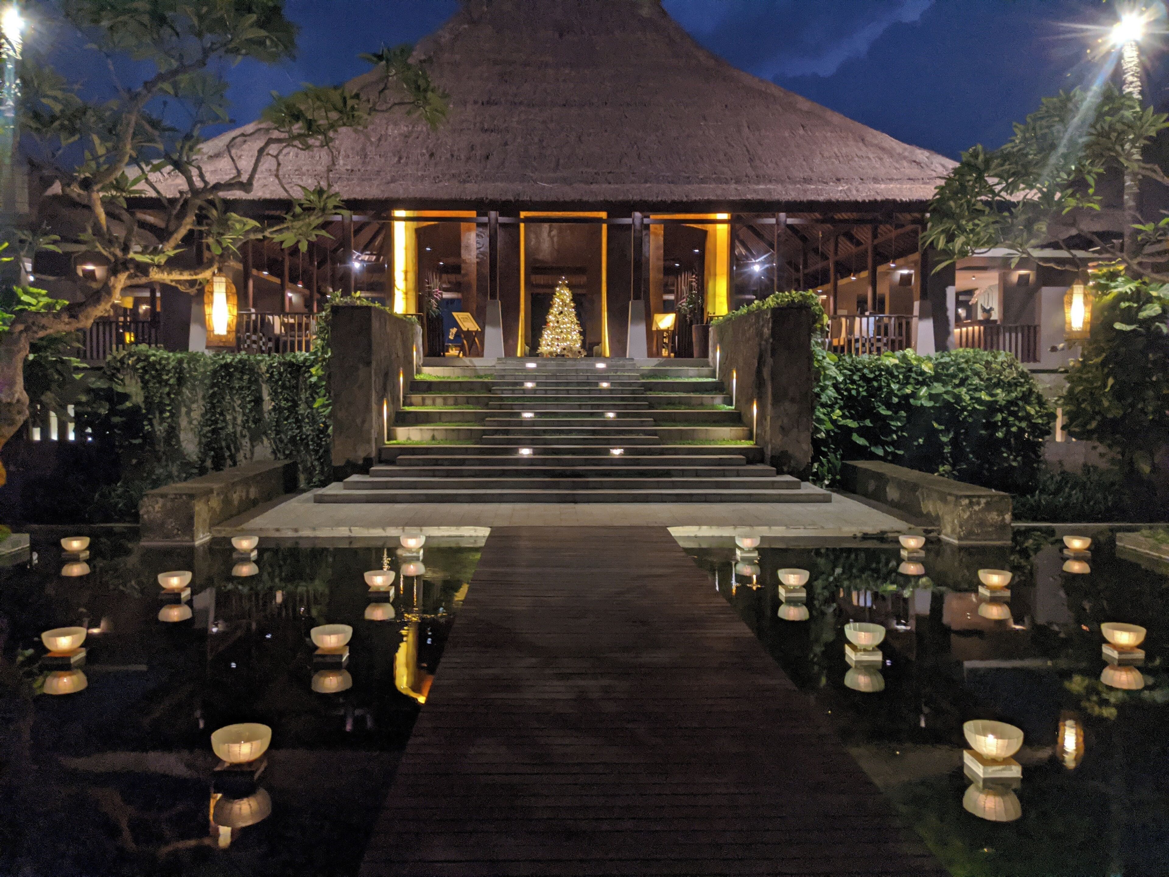 A night view of a resort with a thatched-roof building, a Christmas tree, and a reflective water feature with candles.