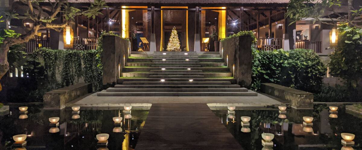 A night view of a resort with a thatched-roof building, a Christmas tree, and a reflective water feature with candles.