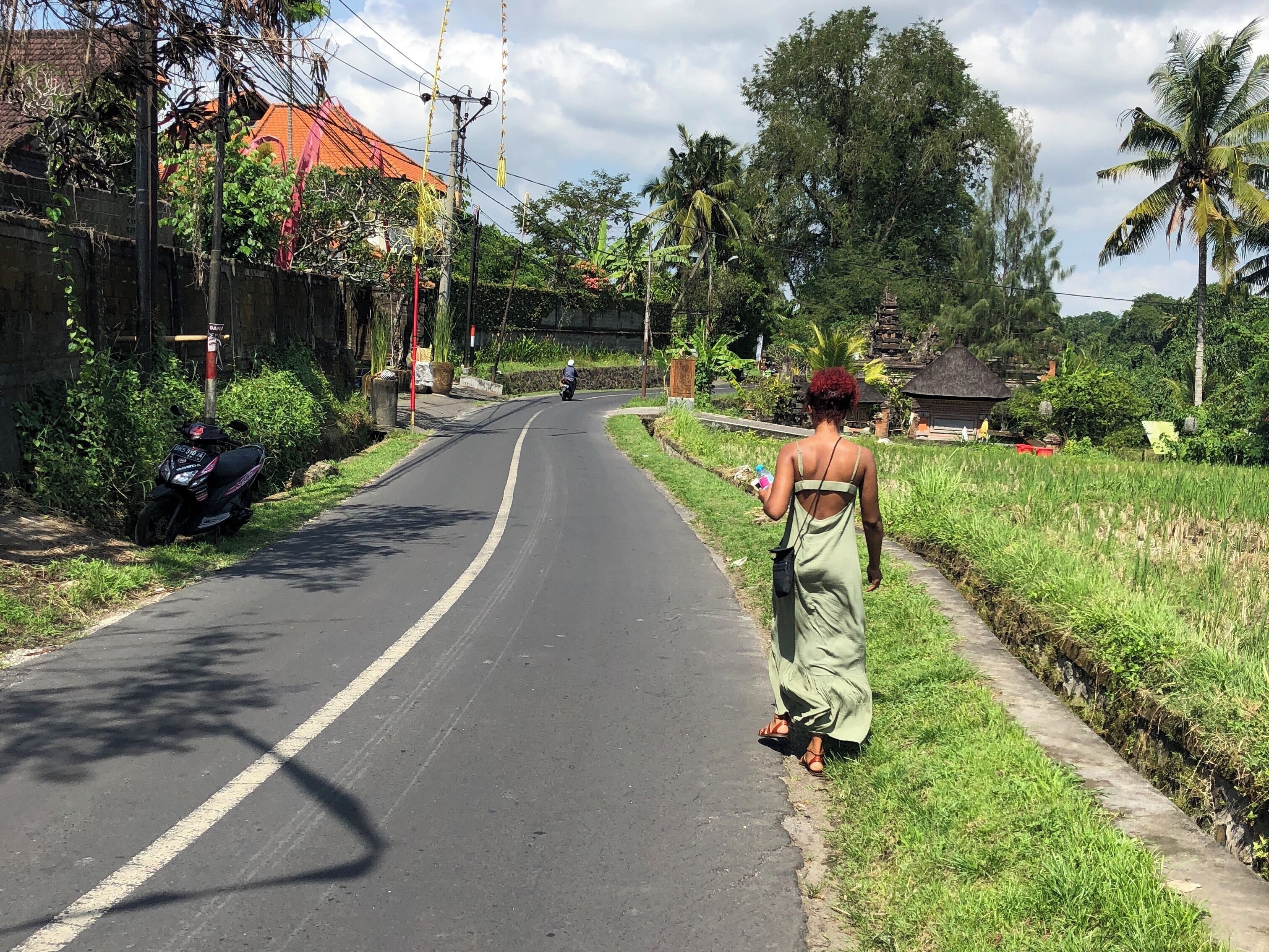 A rural road flanked by lush greenery, a motorbike parked on the side, and a woman walking away from the camera.