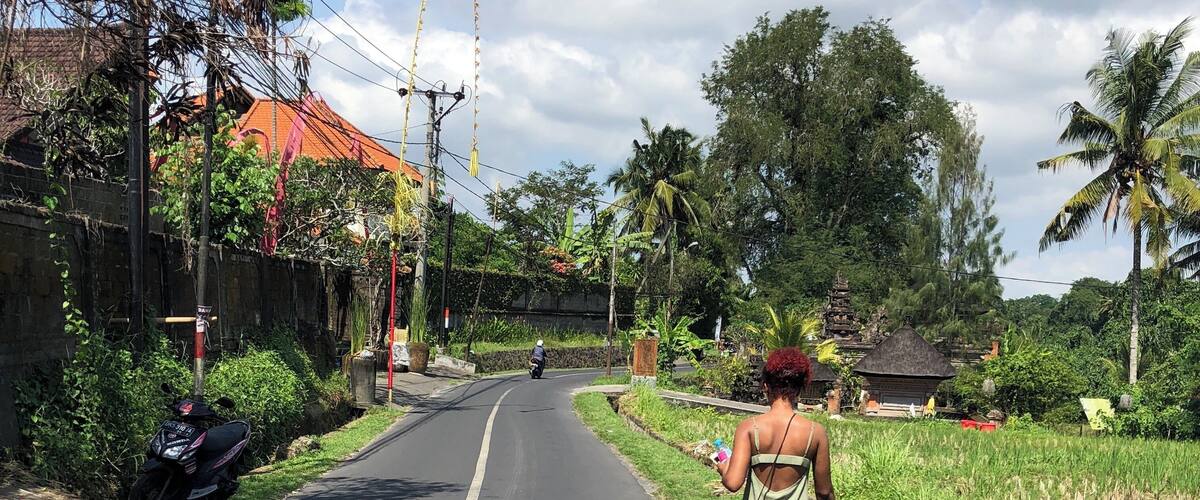 A rural road flanked by lush greenery, a motorbike parked on the side, and a woman walking away from the camera.
