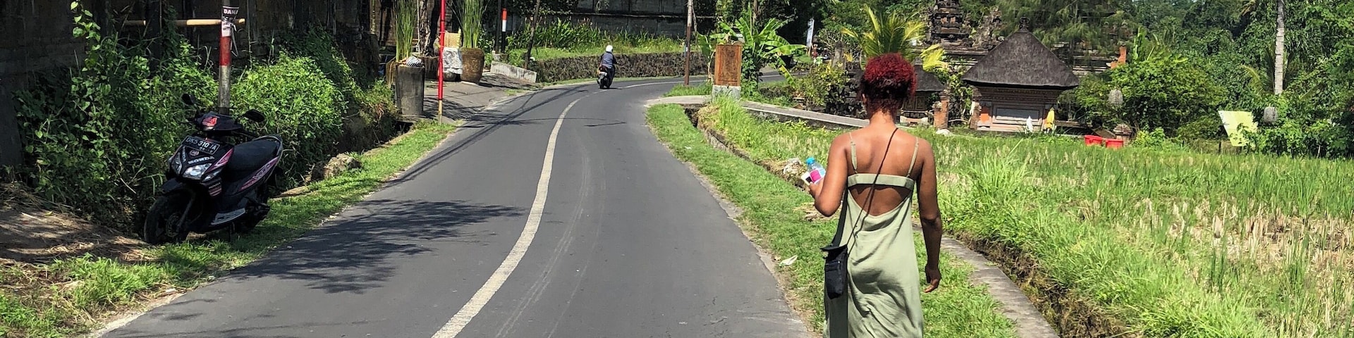 A rural road flanked by lush greenery, a motorbike parked on the side, and a woman walking away from the camera.
