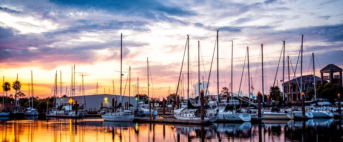 A marina at sunset with multiple sailboats docked, reflecting in calm waters.