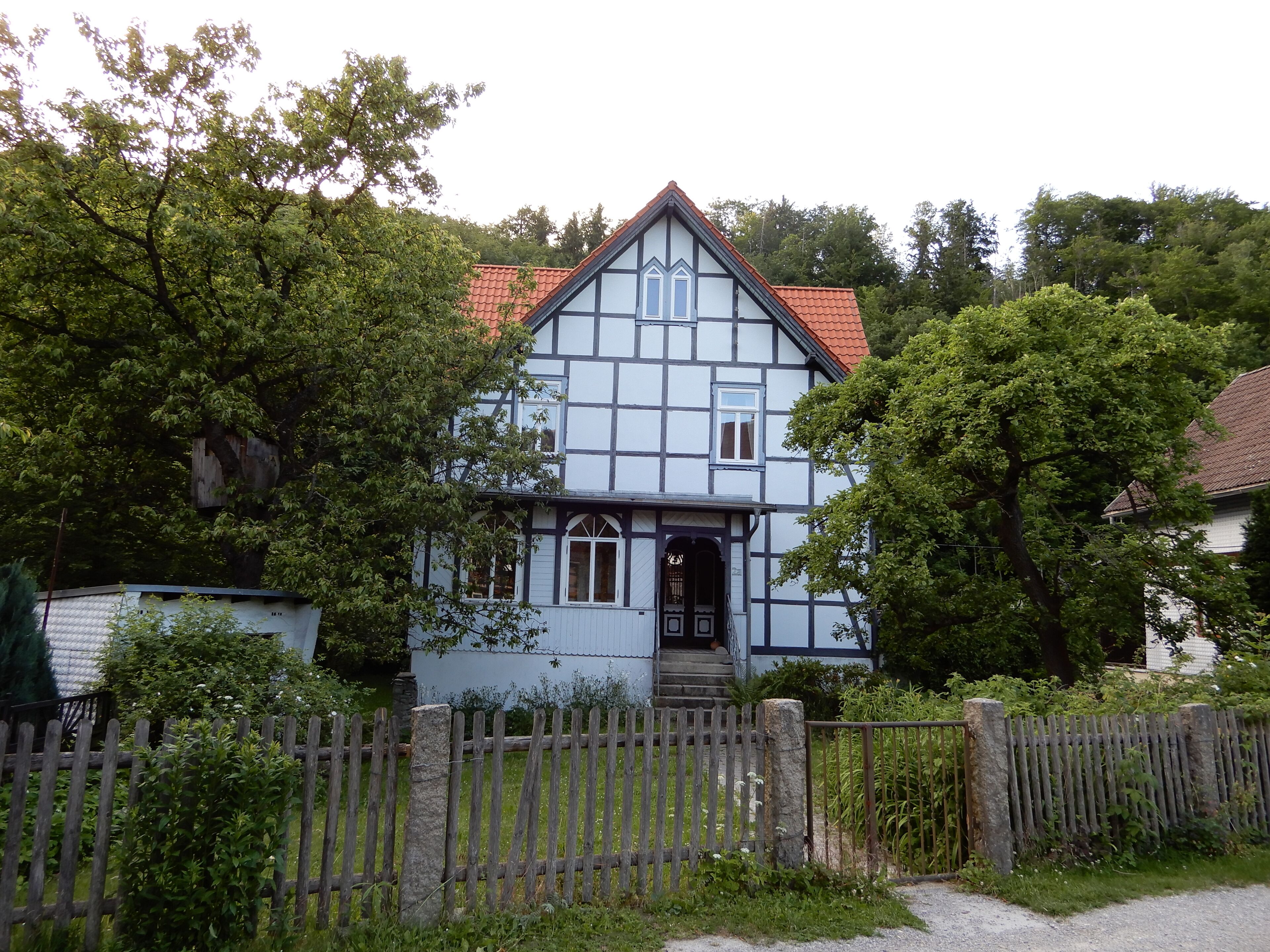 A traditional half-timbered house with a red-tiled roof, surrounded by lush greenery and a wooden fence.