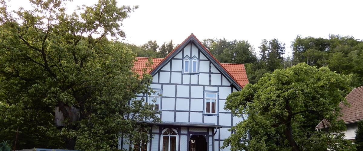 A traditional half-timbered house with a red-tiled roof, surrounded by lush greenery and a wooden fence.