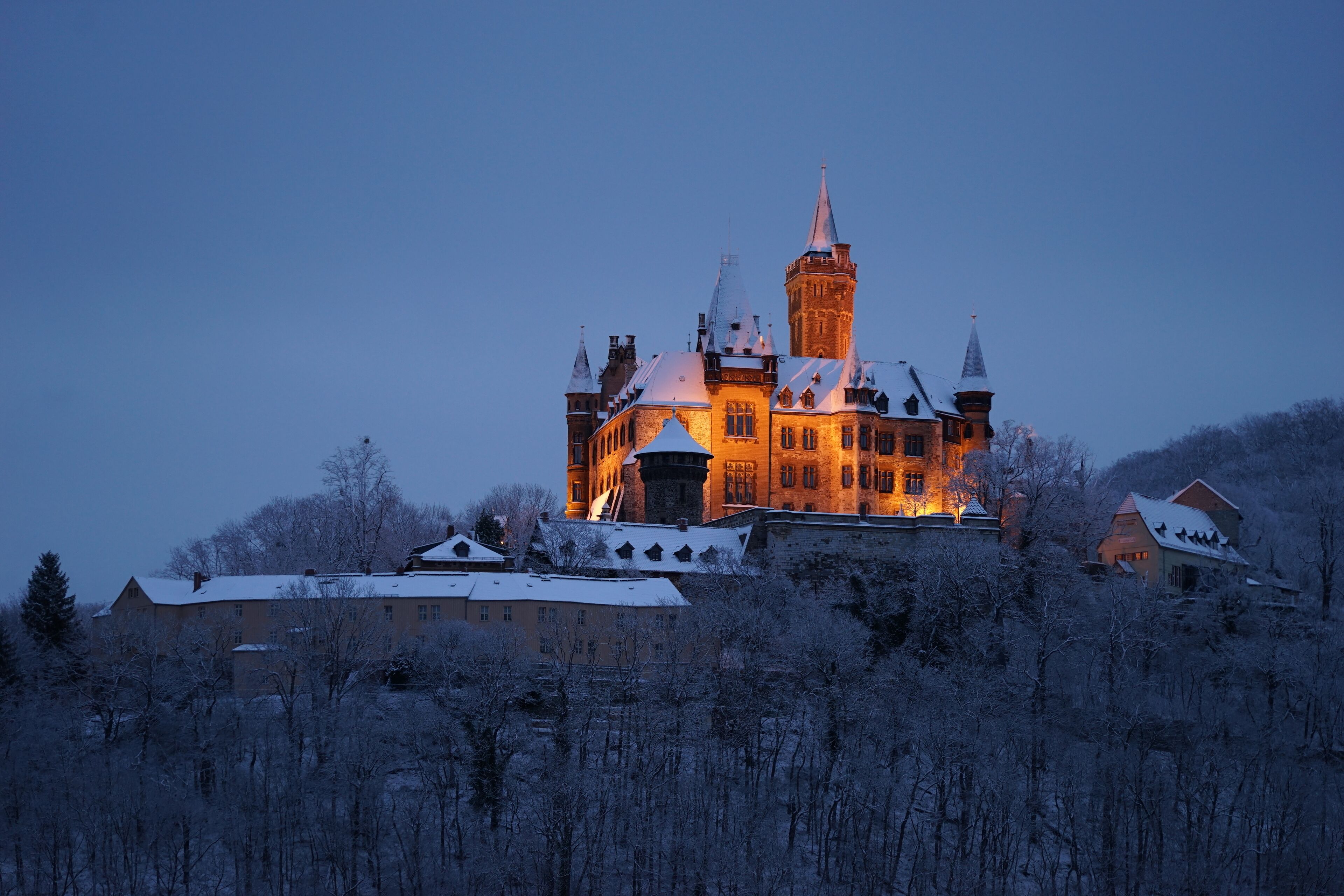 A castle with multiple towers and a prominent central tower, surrounded by snow-covered trees and buildings.