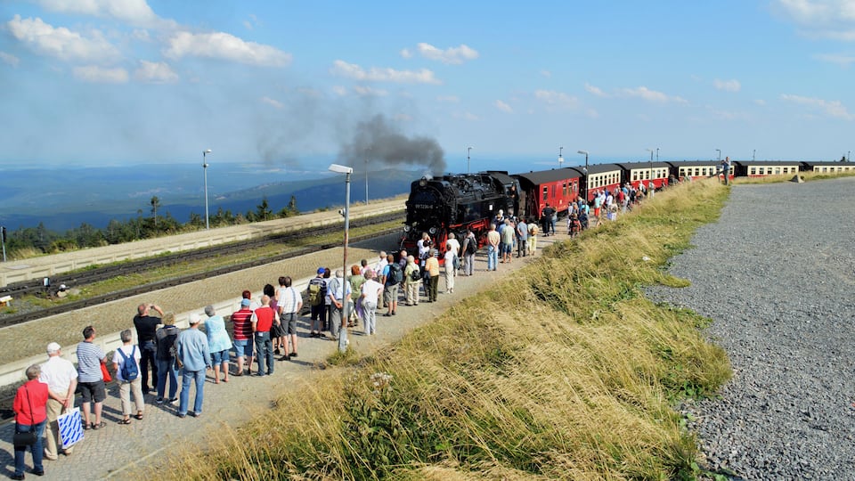 A steam locomotive pulling a red passenger train with passengers standing on the tracks.