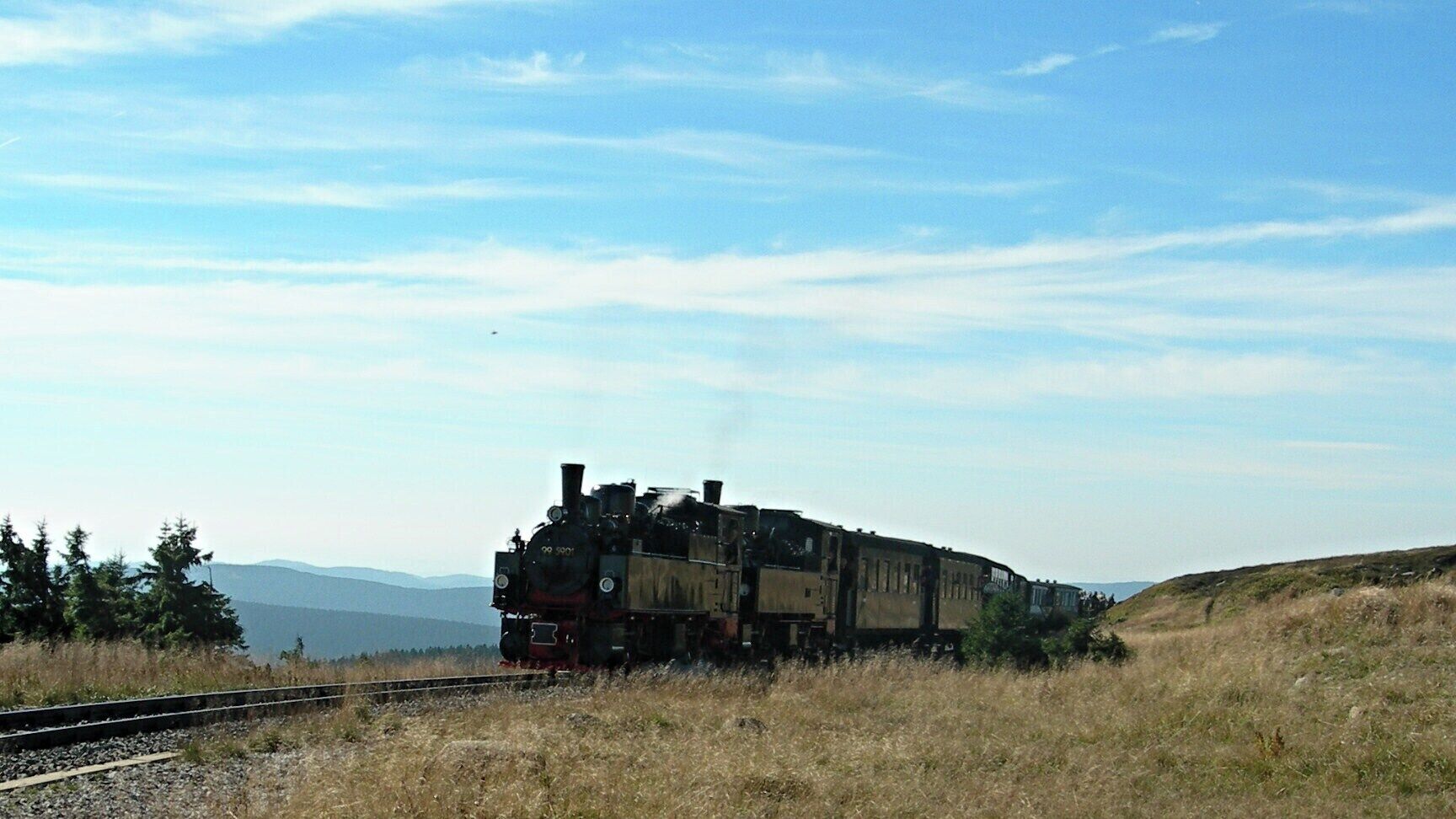 A steam locomotive on a railway track in a rural area with mountains in the background.