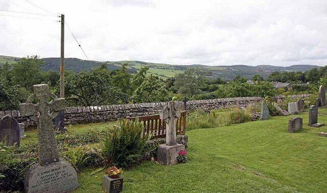 A rural cemetery with stone crosses and headstones, surrounded by lush greenery and rolling hills.