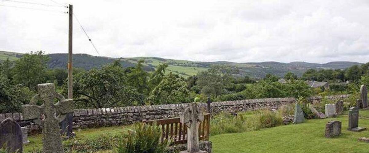 A rural cemetery with stone crosses and headstones, surrounded by lush greenery and rolling hills.