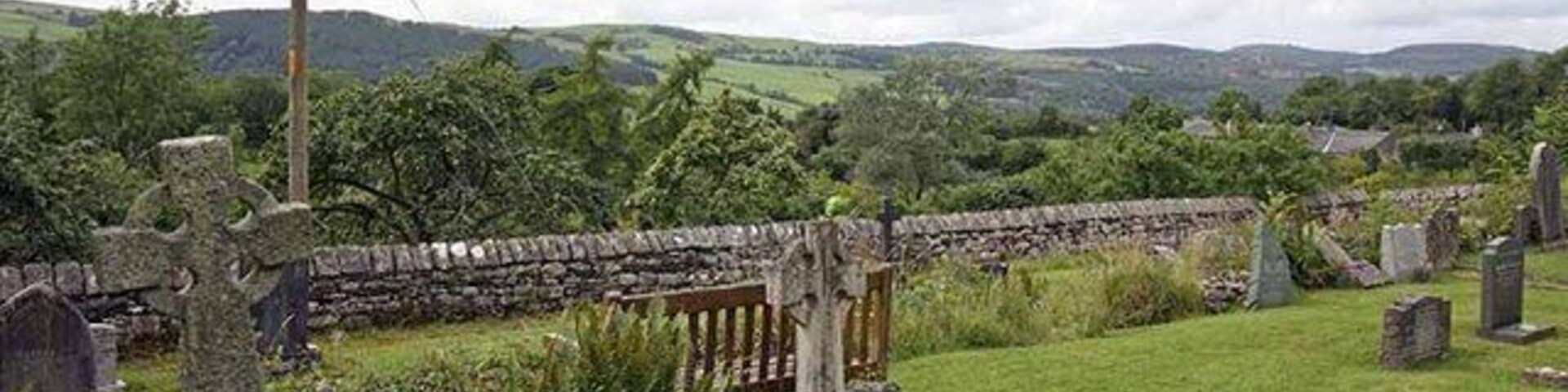 A rural cemetery with stone crosses and headstones, surrounded by lush greenery and rolling hills.