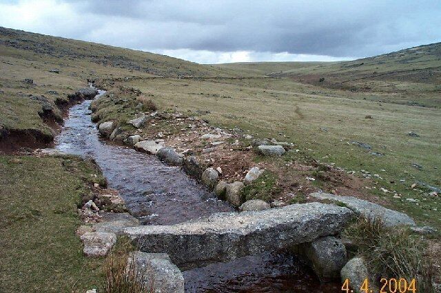 A narrow stream flowing through a grassy valley with rocky banks and a cloudy sky.