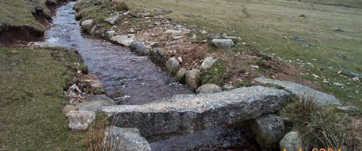 A narrow stream flowing through a grassy valley with rocky banks and a cloudy sky.