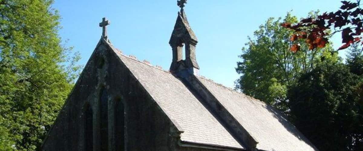 A small stone church with a bell tower and a cross on top, surrounded by greenery.