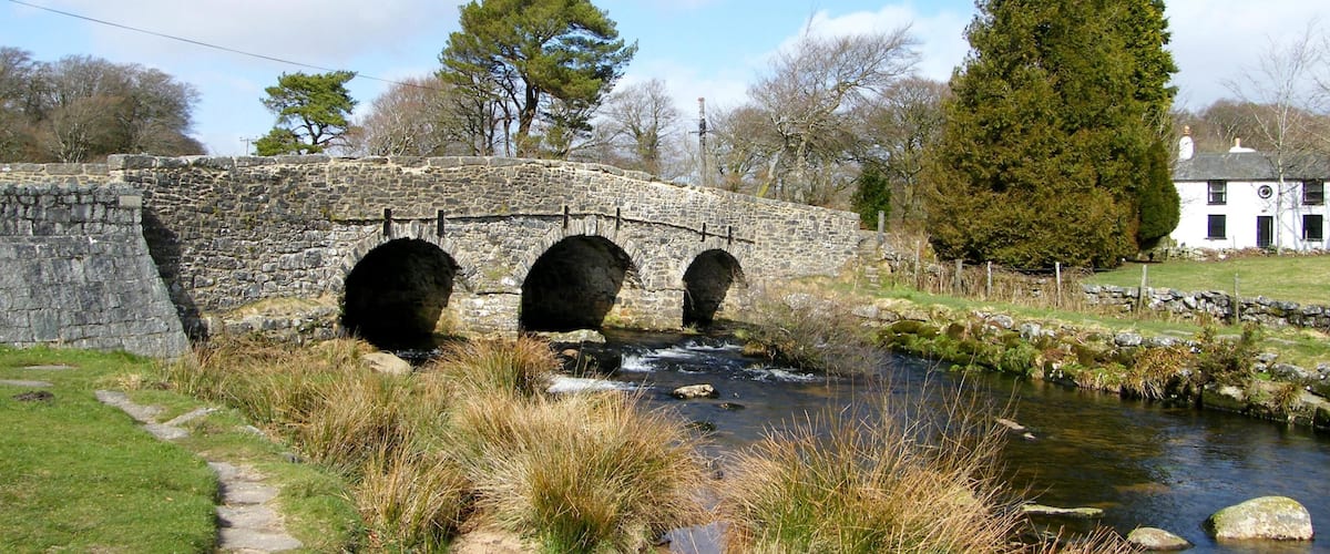 A stone bridge over a stream, surrounded by grass, trees, and a white house.