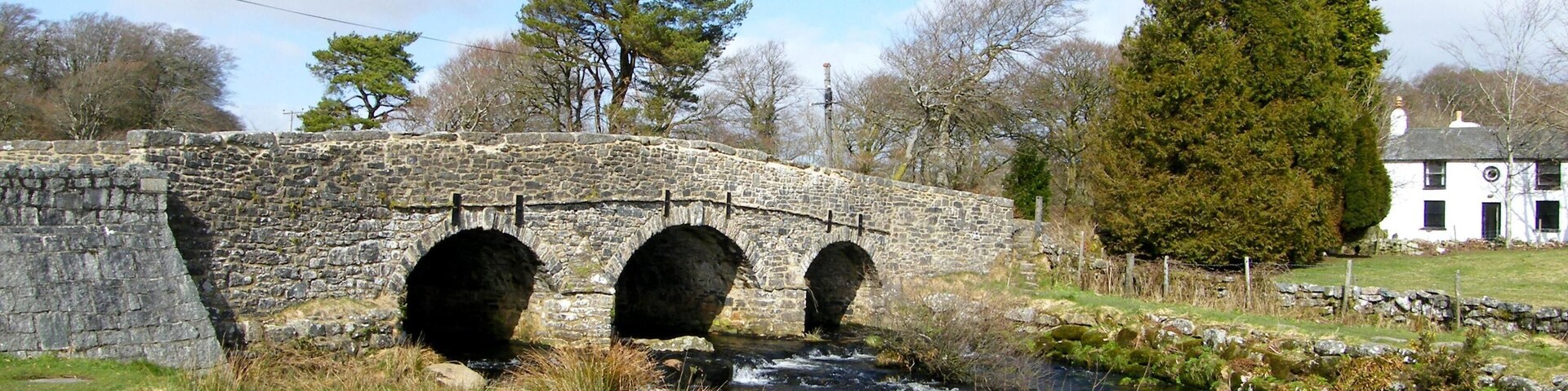 Un puente de piedra sobre un arroyo, rodeado de hierba, árboles y una casa blanca.