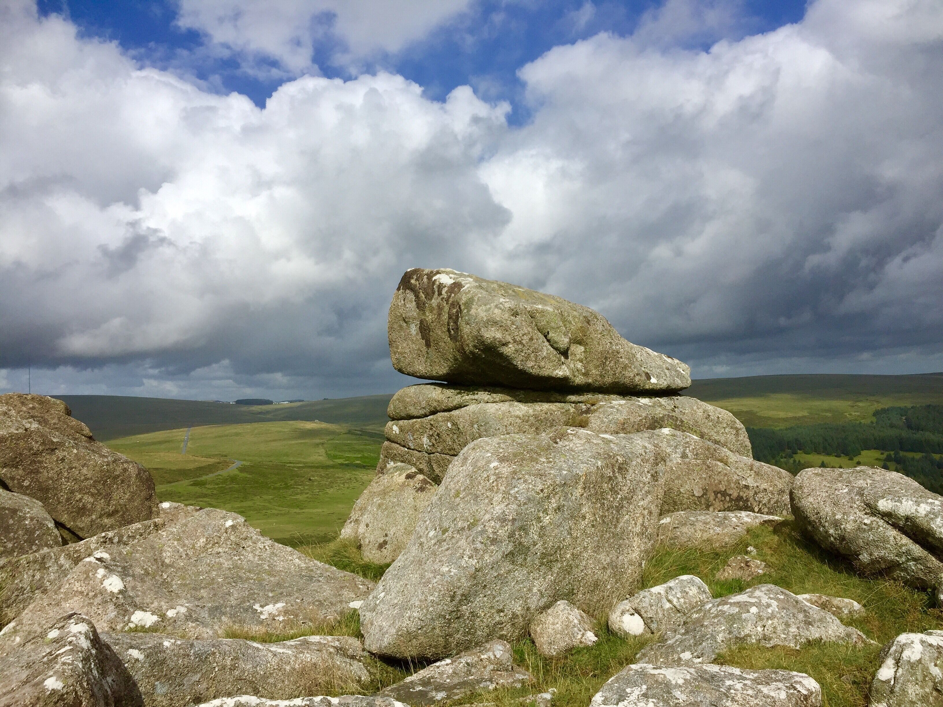 A rocky landscape with a prominent rock formation in the foreground and a vast green valley under a cloudy sky.