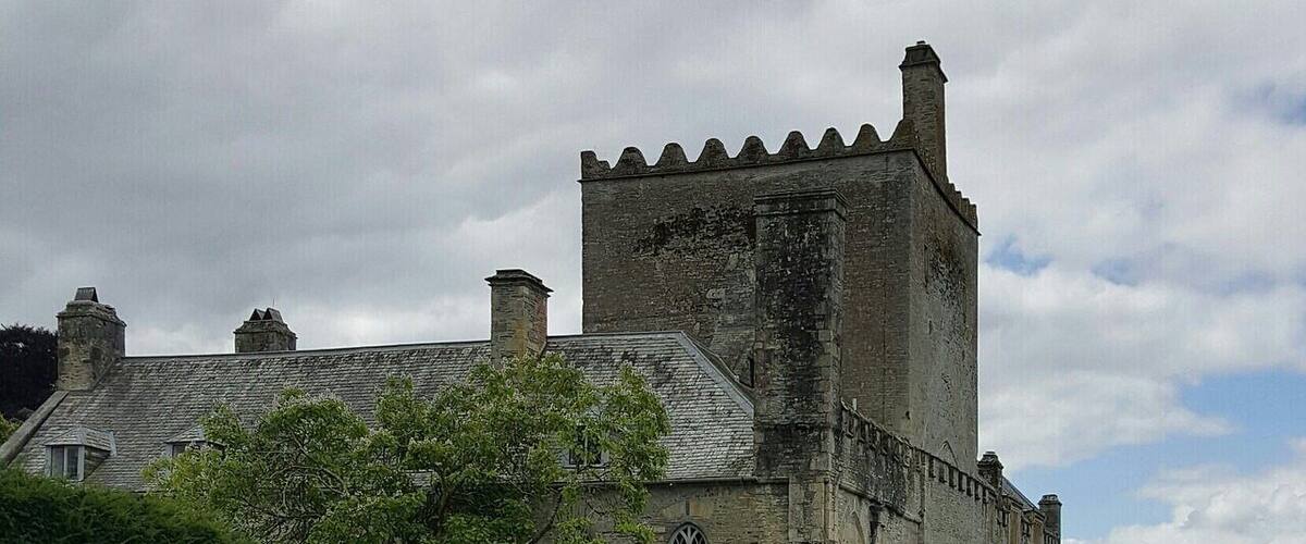 A historic stone castle with battlements, surrounded by lush greenery and a gravel path.