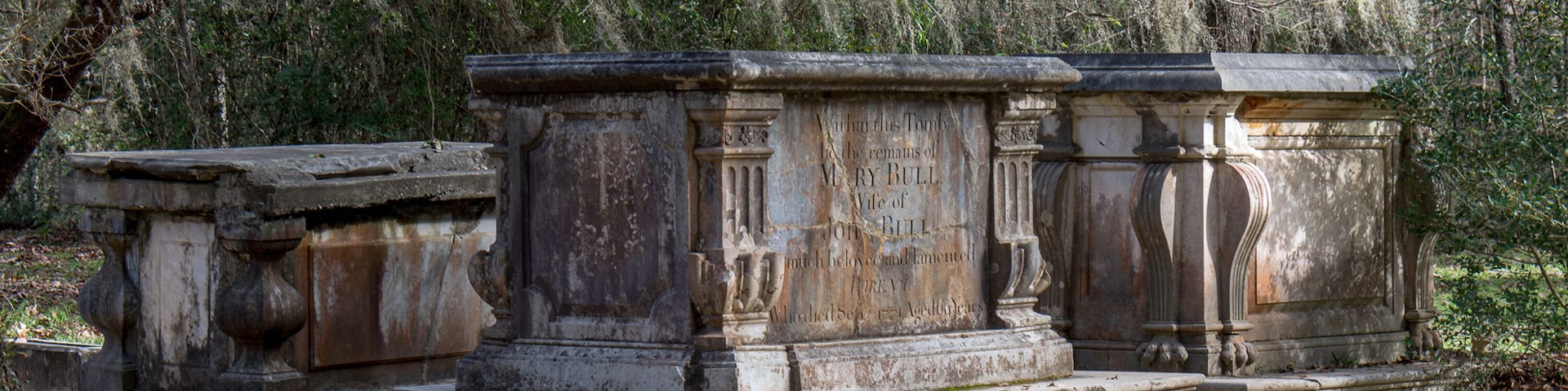 Um monumento de pedra desgastada com inscrições, rodeado por musgo e árvores numa área arborizada.