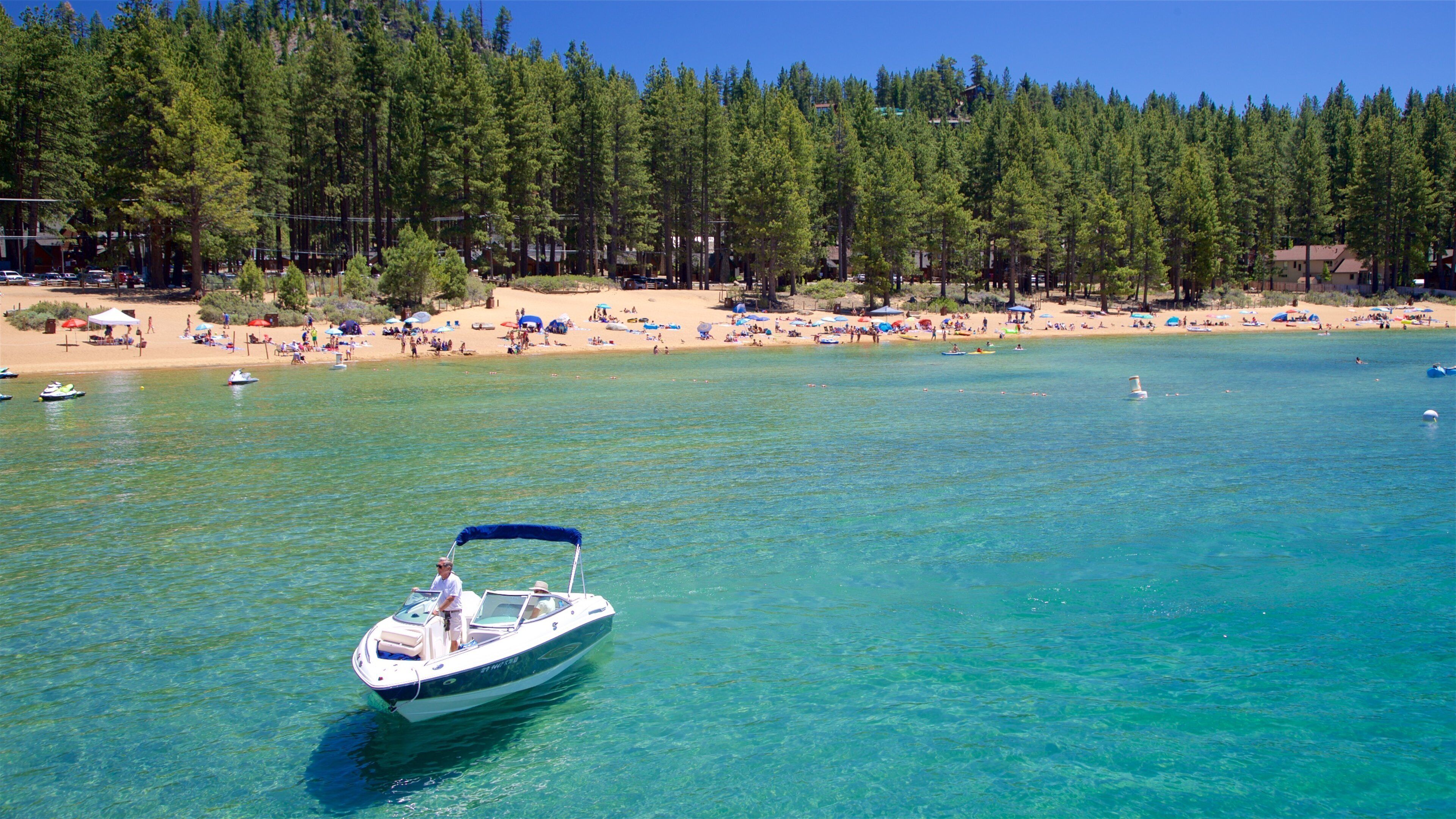 A clear blue lake with a boat in the foreground and a beach with people and umbrellas in the background.