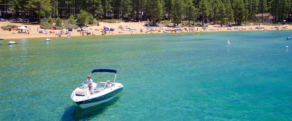 A clear blue lake with a boat in the foreground and a beach with people and umbrellas in the background.