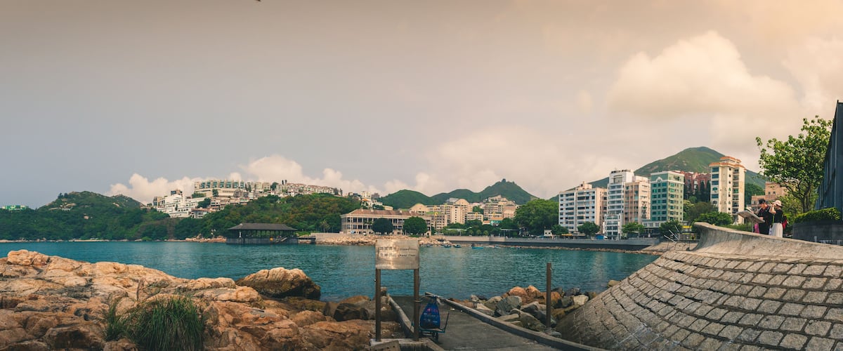 A coastal cityscape with rocky shores, a pier, and buildings on a hillside.